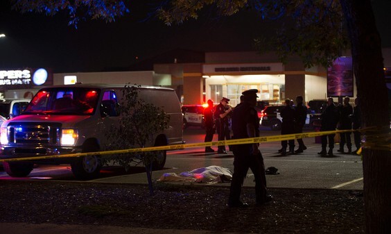 A photo of the crime scene at night, roped off with yellow crime scene tape. A body on the ground is covered by a white tarp; a group of first responders are gathered in the background and a police officer walks past in the foreground.