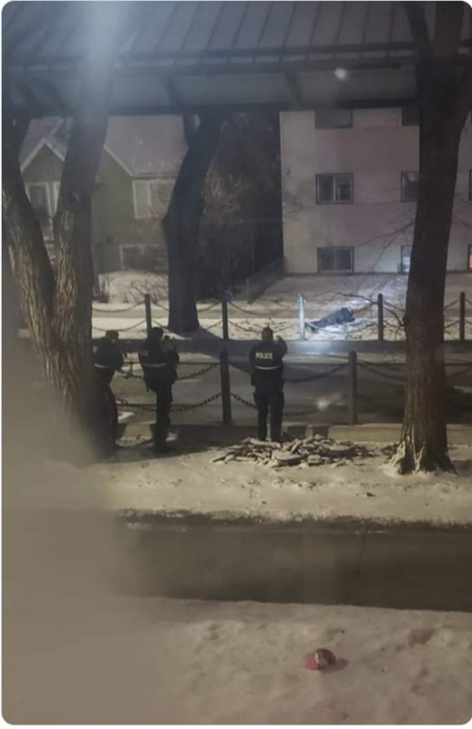 A photo taken from an apartment building shows three EPS officers pointing weapons at a body lying prone in the snow across the street, in front of a low-rise apartment block.
