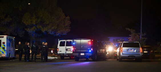 A photo of the crime scene at night, showing several police vans, an ambulance, and a group of first responders (police and paramedics) gathered nearby. Behind them, a white tarp is laid over a body on the ground.