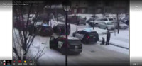 Bystander video recorded from a balcony of the shooting - quality is poor but it shows at least five police cruisers parked in a row in a snowy parking lot. One officer is ducked behind the rear cruiser with a person in handcuffs kneeling on the ground. Much of the scene is obscured by trees but officers can be seen running between vehicles, and numerous gun shots are heard.