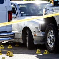 A photo of a white sedan with multiple bullet holes in the windshield and the front bumper compressed into the front of a black van. Numerous yellow evidence markers and police tape surround the scene.