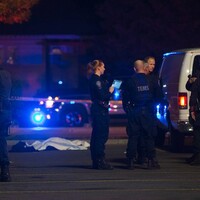 A photo of the crime scene at night, with a white tarp laid over a body on the ground. In the foreground, a group of first responders including police, and TEMS (Tactical Emergency Medical Support) personnel are gathered, next to several police vans.