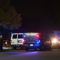 A photo of the crime scene at night, showing several police vans, an ambulance, and a group of first responders (police and paramedics) gathered nearby. Behind them, a white tarp is laid over a body on the ground.