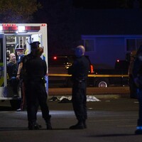 A photo of the crime scene at night. A body covered by a white tarp lies on the ground between a white van and an ambulance. First responders are gathered nearby and in the back of the ambulance.