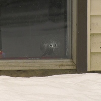A close-up of a basement apartment window with a bullet hole through the glass. A superman figurine sits inside the windowsill.